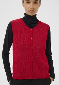 Punainen neulottu hihaton neuletakki V-kaula-aukolla, jossa on resoritekstuuri ja viisi vastaavaa nappia edessä, kerroksittain mustan poolopaidan päällä.