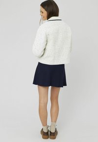 Veste en tweed blanc avec un motif de grille texturé, un col bleu marine, associée à une jupe bleu marine. Chaussures brunes avec des détails en lacets et des chaussettes légères.