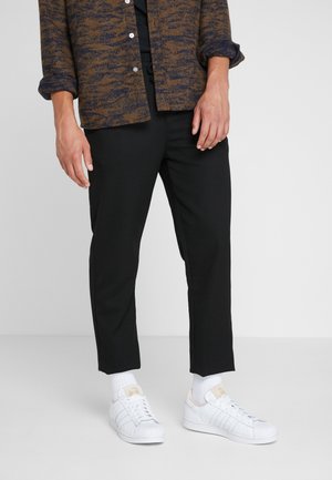 Braun und marineblau gemusterte Button-Up-Hemd über einem schwarzen Outfit, kombiniert mit weißen Sneakern und Socken in Crew-Länge. Glattes Material, lockere Passform.