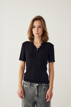Ung kvinde med skulderlangt lyst brunt hår iført en sort kortærmet trøje og grå jeans, stående foran en ensfarvet hvid baggrund.