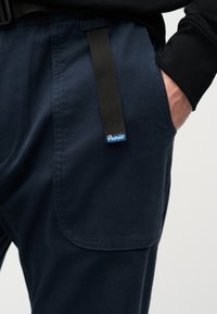 Κοντινό πλάνο σε ναυτικό παντελόνι με μεγάλο πλαϊνό τσέπη, μαύρη θηλιά ζώνης και χέρι που βρίσκεται εν μέρει μέσα στην τσέπη.