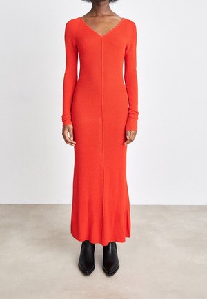 Kobieta ubrana w długą, dopasowaną pomarańczową sukienkę maxi z długim rękawem oraz czarne botki z ostrym noskiem, stojąca na tle jasnej, jednolitej ściany.