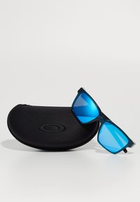 Gafas de sol con tinte azul y un marco de plástico negro, lentes angulados y un estuche duro negro con un logo en relieve.