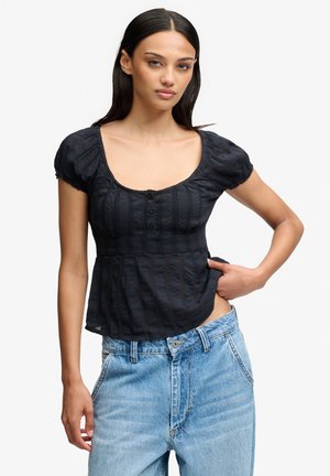 Femme aux longs cheveux foncés portant un chemisier noir à manches courtes et un jean bleu clair, posant avec une main sur la hanche devant un fond uni.