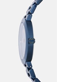 Slim temno modra ura, gledana s strani, prikazuje ohišje ure, krono z logotipom AX in povezano kovinsko zapestnico.