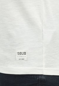 Hvid kortærmet T-shirt lavet af blødt stof, med et firkantet mærke, der har "SOLID DENMARK EST.1999" på nederste venstre side.