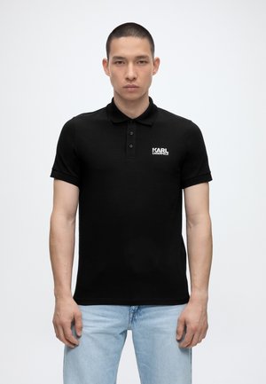 Mladý muž s krátkymi vlasmi, ktorý má na sebe čierne polo tričko Karl Lagerfeld a svetlomodré džínsy, stojí pred jednoduchým bielym pozadím.