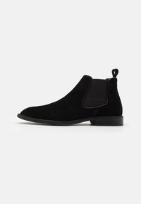 Bottine Chelsea en suède noir avec des panneaux élastiques sur les côtés, bout rond et poignée. Semelle slim avec un léger talon, texture lisse.