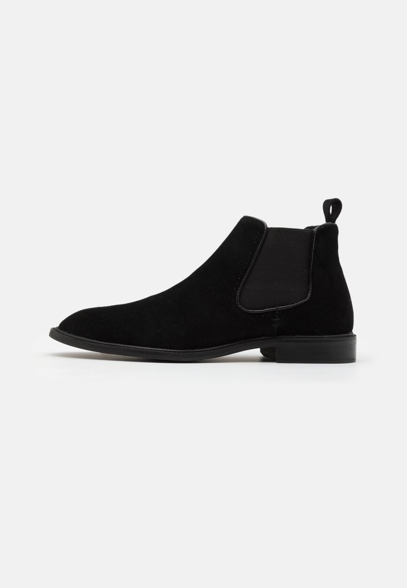 Bottine Chelsea en suède noir avec des panneaux élastiques sur les côtés, bout rond et poignée. Semelle slim avec un léger talon, texture lisse.