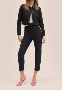 Zwarte cropped jas met knopen, fitted zwarte broeken met zijknopen en roze gestrapte hoge hakken. Glad textuur.