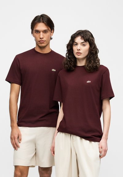 Мъж и жена носят съвпадащи тъмно бордо тениски Lacoste и светлобежови долнища, изправени пред обикновен светъл фон.