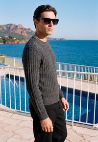 Un homme portant des lunettes de soleil noires et un pull gris en maille torsadée se tient près d'une piscine clôturée avec la mer et les montagnes en arrière-plan.