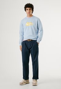 Lichtblauwe sweatshirts met gele tekst, marineblauwe broek en beige sneakers. De sweatshirt heeft een ronde halslijn en ribgebreide manchetten.