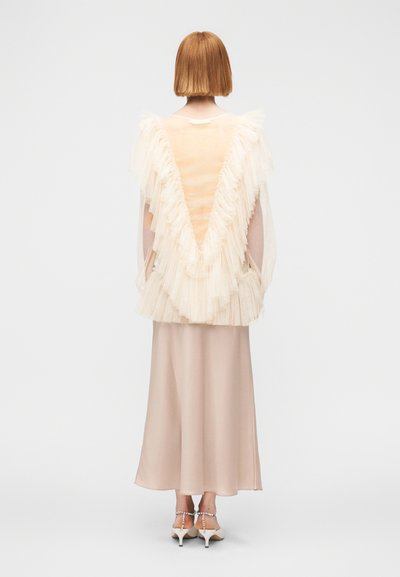 Αέρινο κρεμ τοπ με στρώσεις τούλινης βολάν, μπεζ σατέν φούστα και λευκά ψηλοτάκουνα σανδάλια με λουράκια. Πλάγια όψη από πίσω.