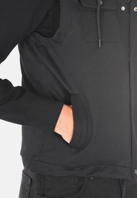 Giacca nera con polsini a costine, tessuto texturizzato e chiusura con zip; presenta una tasca laterale integrata nel design.