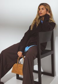 Sort oversized skjorte, lyseblå undertrøje, brune stribede bukser og en brun ruskinds håndtaske. Sittende på en sort stol.