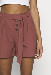 Højtaljede shorts i en støvet rosa farve, lavet af et tekstureret stof, med et bundet taljebånd og tre dekorative knapper.