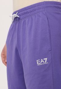 Violetiniai medvilniniai džinsai su elastine juosta, turintys baltą EA7 Emporio Armani logotipą apatinėje dalyje ir šonines kišenes.