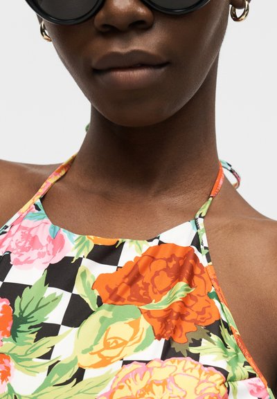 Nærbilde av en person med svarte runde solbriller, gullringleer, og en blomstrete haltertop med oransje, rosa og gule blomster på rutete bakgrunn.