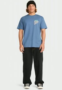Hombre con gorro negro, camiseta azul de Billabong, pantalones negros sueltos y zapatillas en blanco y negro, de pie contra un fondo sencillo.