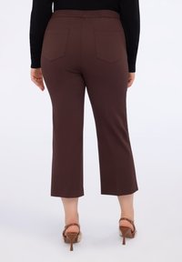 Persoană îmbrăcată într-o pereche de pantaloni scurți maro, un top negru cu mânecă lungă și sandale cu toc înalt maro, stând cu spatele pe un fundal alb.