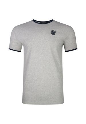 Mężczyzna z tatuażami na ramionach i szyi, w dopasowanym szarym t-shircie, luźnych czarnych spodniach, białych tenisówkach i bransoletkach, stojący na szarym tle.