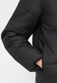 Svart puffjakke med polstret design, med en avslappet passform og en sidelomme. Materialet ser glatt og litt glinsende ut.