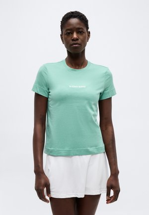 Γυναίκα που φορά ένα ανοιχτοπράσινο t-shirt Björn Borg και λευκά σορτς, στέκεται μπροστά σε απλό γκρι φόντο.