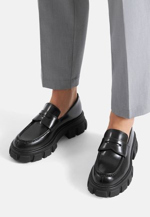 Mocasines de cuero negro con suela gruesa de goma. Presentan una correa decorativa sobre el empeine y una costura detallada a lo largo de los bordes.