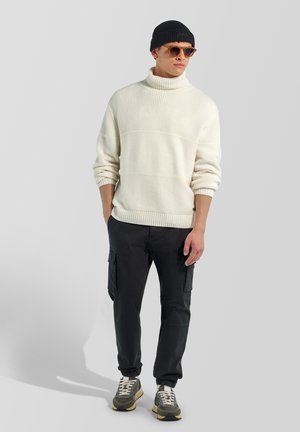 Giovane uomo che indossa un maglione a collo alto bianco, pantaloni cargo neri, sneakers, berretto nero e occhiali da sole, in piedi con le mani in tasca.