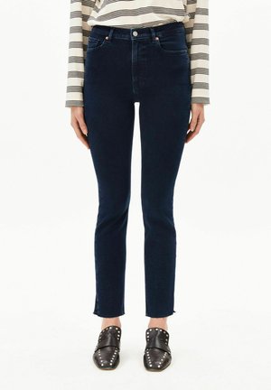 Person trägt dunkelblaue schmale Jeans, ein beiges langärmeliges Hemd mit schwarzen Streifen und schwarze Nieten-Slipper.