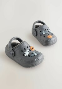 Sabots gris avec un intérieur doux et duveteux, ornés de décorations en plastique : une momie, une citrouille, une chauve-souris et un fantôme sur le dessus.