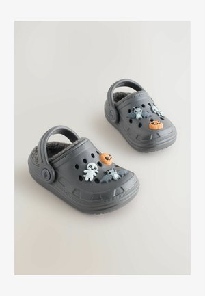 Šedé dřeváky s měkkým, huňatým interiérem, zdobené plastovými přívěsky: mumie, dýně, netopýr a duch na horní části.