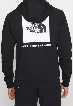 Schwarzer Hoodie mit weißem quadratischem Logo "THE NORTH FACE" und Slogan "NEVER STOP EXPLORING" auf dem Rücken, getragen von einer Person, die wegschaut.