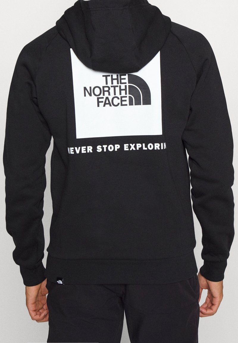 Sweat à capuche noir avec un logo carré blanc indiquant "THE NORTH FACE" et le slogan "NEVER STOP EXPLORING" au dos, porté par une personne vue de dos.