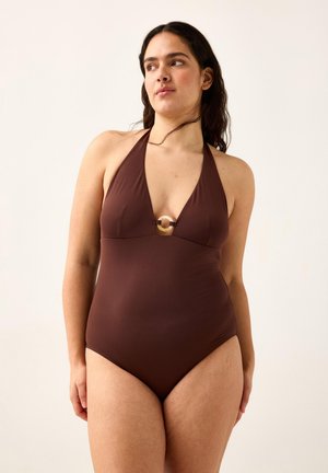 Kvinde med langt mørkt hår iført en brun halterneck badedragt med en cirkulær gulddetalje ved brystet, stående foran en ensfarvet baggrund.