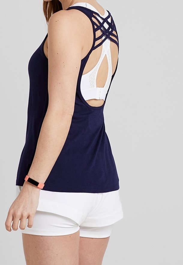 Femme portant un débardeur bleu marine avec dos croisé, un soutien-gorge de sport blanc, un short blanc et un bracelet connecté corail au poignet gauche.