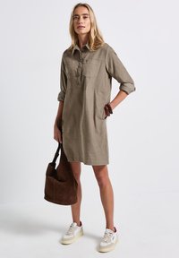 Olivgrön linnedress med uppvikt ärm, knappfront och två bröstfickor, i kombination med en brun mockaväska och vita sneakers.