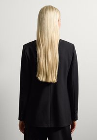 Blazer nero sartoriale con un design strutturato, con maniche lunghe e polsini abbottonati, visto da dietro su uno sfondo neutro.