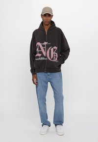Haut de sweatshirt zippé noir avec un grand texte graphique rose. Associé à un jean bleu clair et des chaussures blanches, avec une casquette beige.