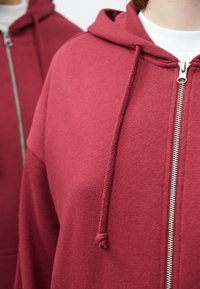 Bordó cipzáras kapucnis pulóver, húzózsinórral ellátott kapucnival, ezüst cipzárral és puha, texturált anyagból. Laza stílust hangsúlyoz.