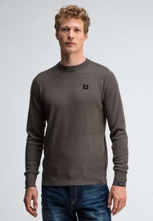 Junger Mann mit lockigem Haar, der einen strukturierten braunen Pullover und blaue Jeans trägt, vor einem schlichten weißen Hintergrund steht.