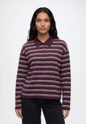 Vrouw met lang donker haar die een maroon- en wit gestreept polo met lange mouwen en zwarte broek draagt, staand tegen een effen achtergrond.