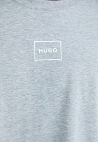 Grå bomull T-skjorte med en enkel hvit rektangulær logo med teksten "HUGO" sentrert på brystet. Myk tekstur, rund hals.