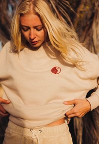 Beżowa bluza z okrągłą czerwoną łatą, miękka w dotyku, oversize'owy krój i okrągły dekolt. Połączona z beżowymi spodniami o teksturowanej fakturze.