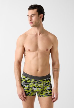 Mężczyzna bez koszulki z ciemnymi włosami, ubrany w zielono-czarno-szare bokserki moro z szarą gumką w pasie z napisem "GUESS", stojący na białym tle.