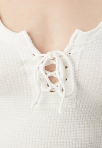 Hvit teksturert topp med V-hals og snøring. Stoffet er ribbet, og snørene er laget av matchende hvitt materiale.