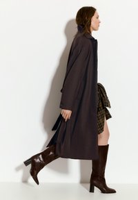 Lang brun frakke lavet af stof, med krave og knaplukning. Kombineret med knæhøje brune støvler med hæl og et ternet skørt.