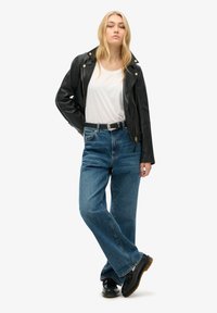 Zwarte lederen motorjack over een witte shirt, gecombineerd met hooggepullde blauwe jeans en zwarte schoenen met een dikke zool.
