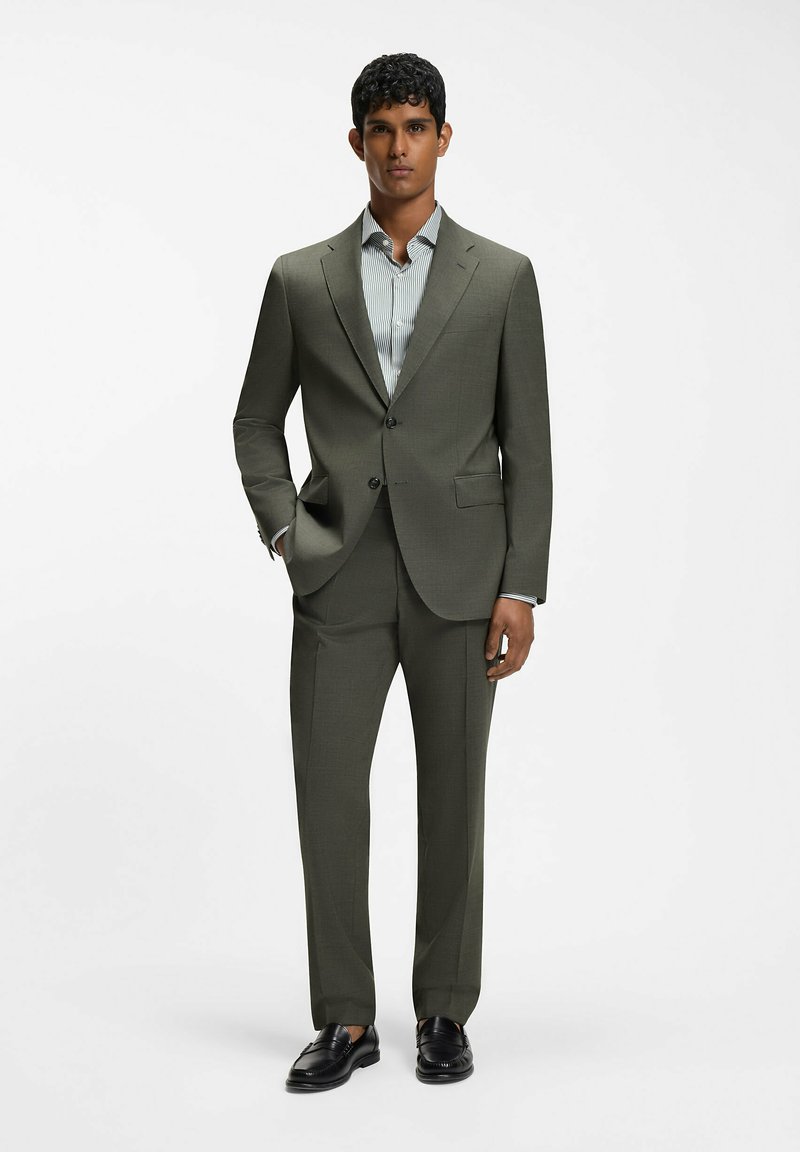 Homme debout portant un costume vert olive avec une chemise à rayures et des chaussures habillées noires sur fond blanc uni.